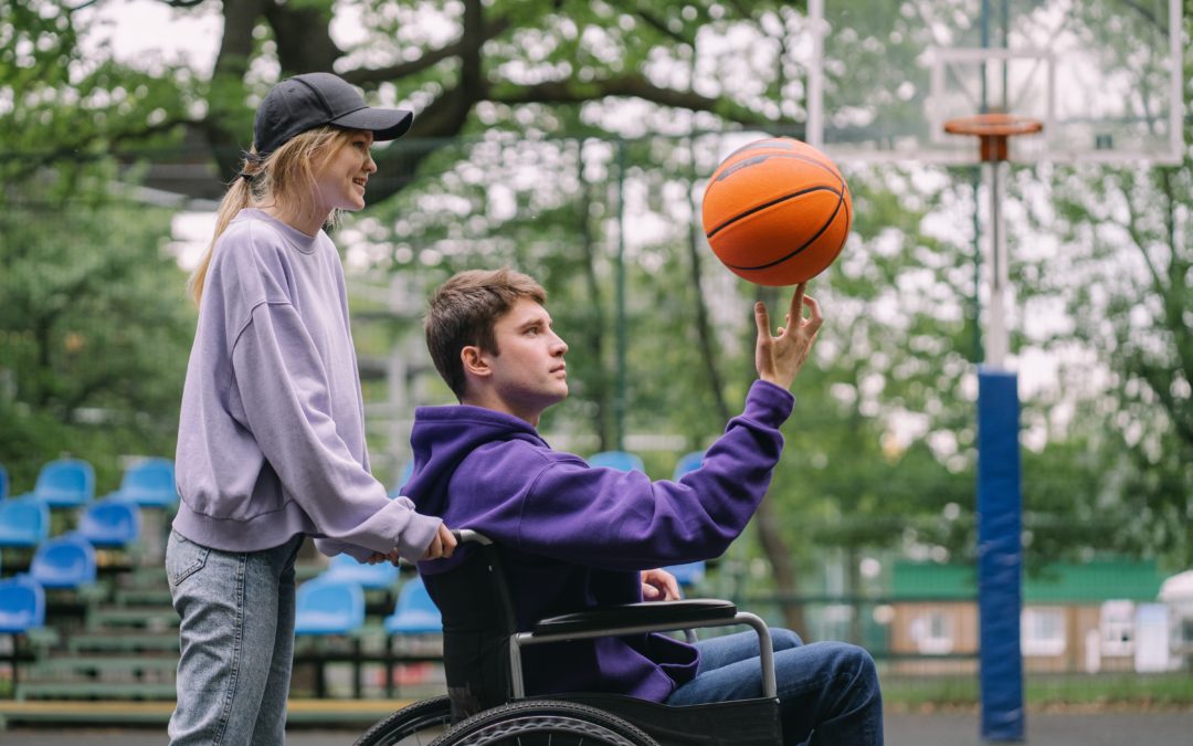 Photo of two teenage students on a basketball court. One male student is in a wheelchair, holding a basketball. The other female student stands behind him with her hands resting on the back of the chair.